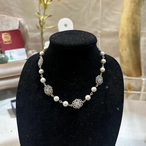 Puede incluir: Un collar dorado con perlas de imitaci&oacute;n y detalles decorativos de filigrana. El collar se exhibe sobre un busto de maniqu&iacute; negro. Las perlas se intercalan con dise&ntilde;os met&aacute;licos calados y ornamentados.