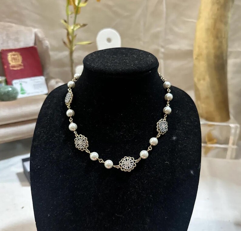 Puede incluir: Un collar dorado con perlas de imitaci&oacute;n y detalles decorativos de filigrana. El collar se exhibe sobre un busto de maniqu&iacute; negro. Las perlas se intercalan con dise&ntilde;os met&aacute;licos calados y ornamentados.
