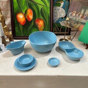May include: A collection of light blue ceramic serving dishes, including a large bowl, a divided dish, a small bowl, a saucer with a bowl, and a gravy boat. The dishes are arranged on a white tablecloth.