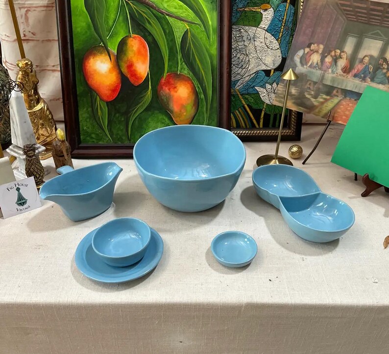 May include: A collection of light blue ceramic serving dishes, including a large bowl, a divided dish, a small bowl, a saucer with a bowl, and a gravy boat. The dishes are arranged on a white tablecloth.