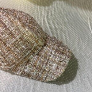 May include: A close-up of a baseball cap with a woven tweed design. The cap features a mix of light pink, beige, and gold threads, with subtle sequin accents. The brim is curved, and the overall texture is slightly rough.