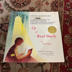 May include: A book cover with a yellow and gold sticker that says "Newbery Award Records" and the title "Up a Road Slowly" by Irene Hunt. The cover features a painting of a person reading a book in a sunlit room.