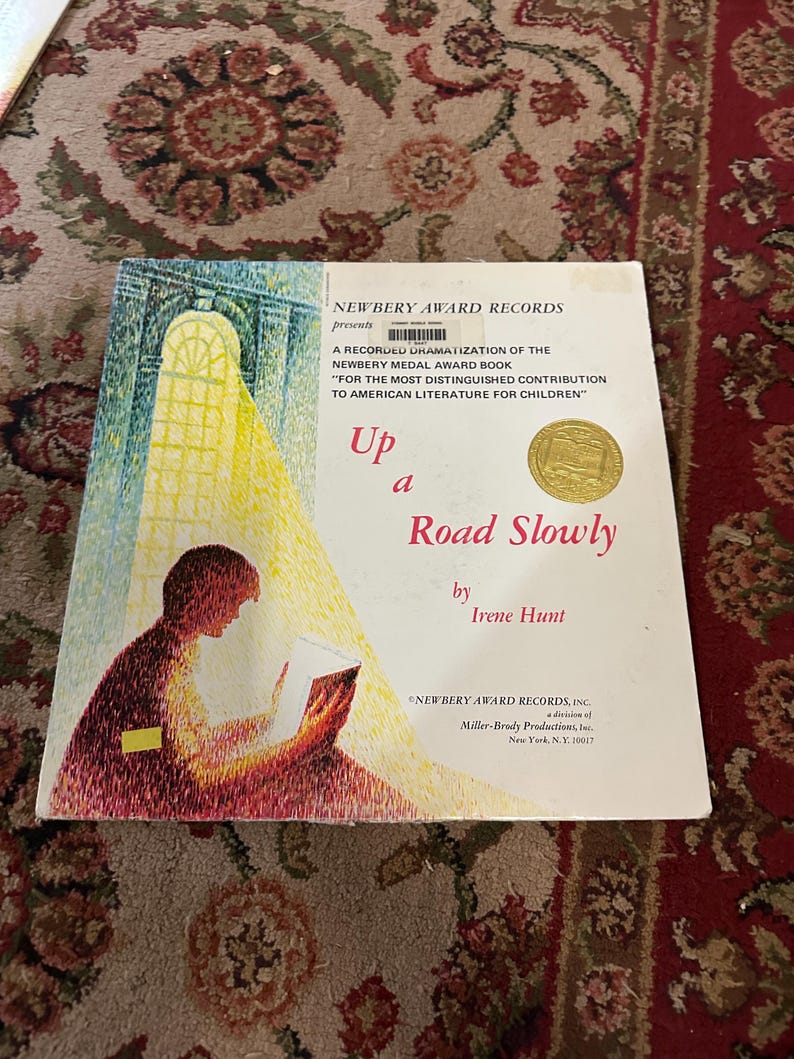 May include: A book cover with a yellow and gold sticker that says "Newbery Award Records" and the title "Up a Road Slowly" by Irene Hunt. The cover features a painting of a person reading a book in a sunlit room.