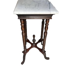 Puede incluir: Una mesa auxiliar cuadrada con un tablero de mármol blanco con vetas grises. La base está construida en madera de color marrón oscuro, con cuatro patas torneadas conectadas por un travesaño en forma de cruz con un remate central.