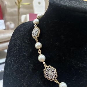 Puede incluir: Un collar dorado con perlas de imitaci&oacute;n y adornos florales calados. El collar se exhibe sobre un fondo negro, resaltando el dise&ntilde;o delicado y el contraste entre el oro y las perlas.