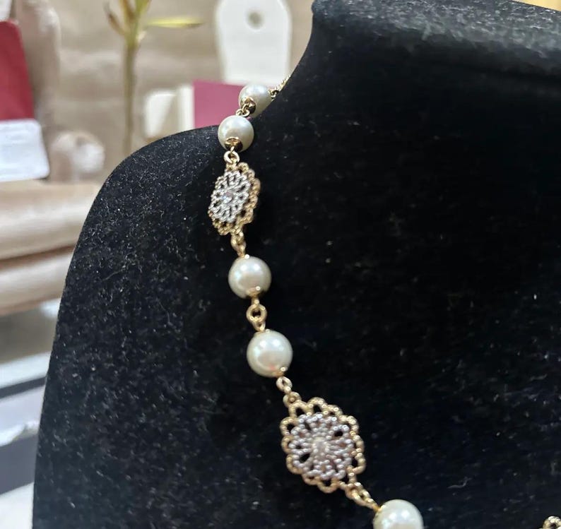 Puede incluir: Un collar dorado con perlas de imitaci&oacute;n y adornos florales calados. El collar se exhibe sobre un fondo negro, resaltando el dise&ntilde;o delicado y el contraste entre el oro y las perlas.