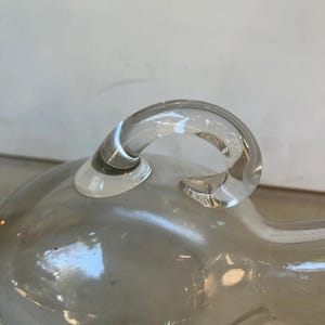 May include: A clear glass bottle with a rounded body and a single handle. The bottle has a smooth, polished surface.