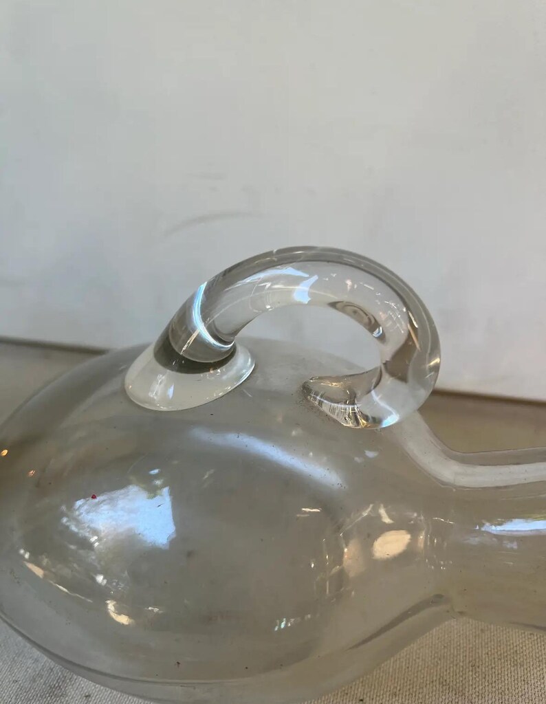 May include: A clear glass bottle with a rounded body and a single handle. The bottle has a smooth, polished surface.