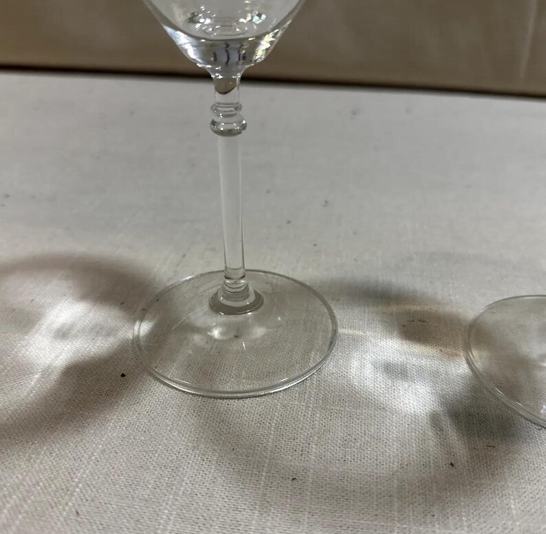 May include: A clear wine glass with a round base and a long, slender stem. The glass is made of clear glass and is partially visible, with the base and stem in focus. The glass is on a white surface.