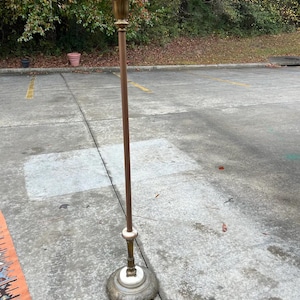 Puede incluir: Una l&aacute;mpara de pie vintage con una parte superior y una base de color lat&oacute;n, un poste marr&oacute;n y un acento blanco. La l&aacute;mpara tiene un dise&ntilde;o ornamentado en la base y una parte superior decorativa. La l&aacute;mpara est&aacute; sobre una superficie de hormig&oacute;n.