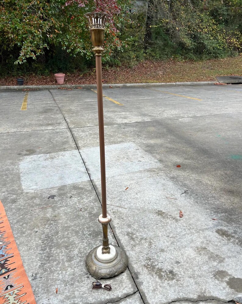 Puede incluir: Una l&aacute;mpara de pie vintage con una parte superior y una base de color lat&oacute;n, un poste marr&oacute;n y un acento blanco. La l&aacute;mpara tiene un dise&ntilde;o ornamentado en la base y una parte superior decorativa. La l&aacute;mpara est&aacute; sobre una superficie de hormig&oacute;n.