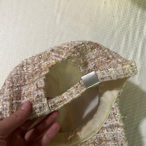 May include: A tweed baseball cap in shades of pink, cream, and gold. The cap features a silver-toned adjustable closure and a cream-colored interior lining. The fabric has a textured appearance.