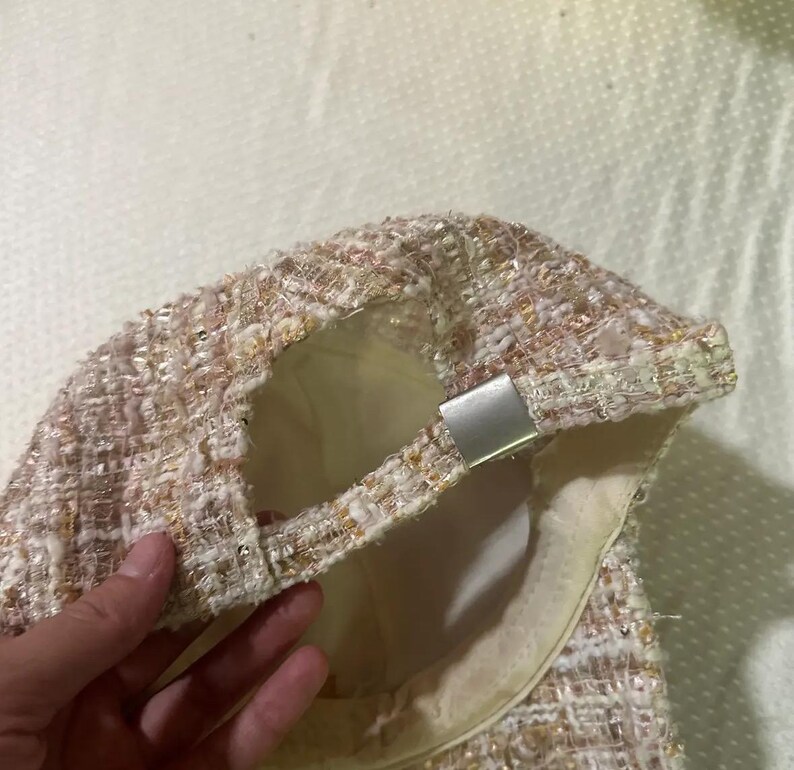May include: A tweed baseball cap in shades of pink, cream, and gold. The cap features a silver-toned adjustable closure and a cream-colored interior lining. The fabric has a textured appearance.