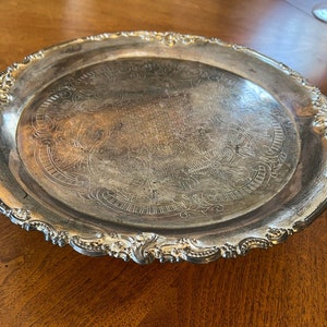 May include: A silver serving tray with an ornate, scalloped edge and an engraved design. The tray is circular and has a raised rim.