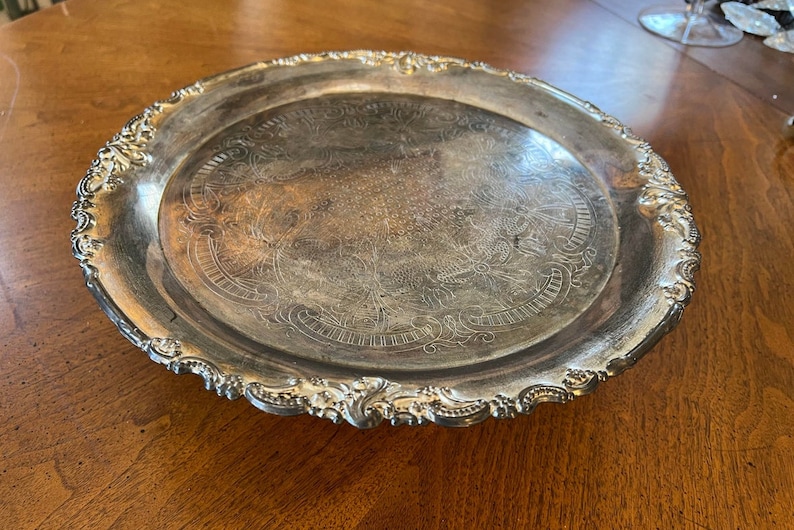 May include: A silver serving tray with an ornate, scalloped edge and an engraved design. The tray is circular and has a raised rim.