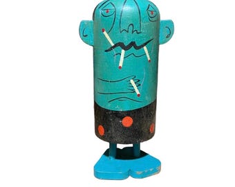 May include: A whimsical, hand-painted wooden figurine. The figure has a turquoise head and body with a black skirt adorned with orange dots. The face has red eyes and a zig-zag mouth, with cigarettes. The figurine stands on blue feet.