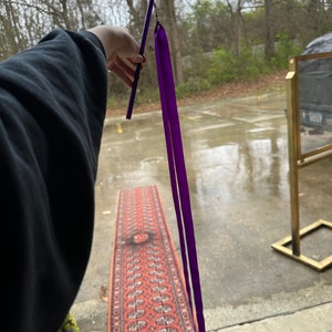May include: A purple ribbon attached to a black stick. The ribbon is flowing in the air. The stick is held by a hand.