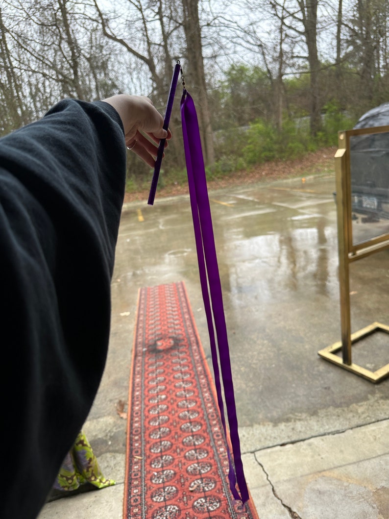 May include: A purple ribbon attached to a black stick. The ribbon is flowing in the air. The stick is held by a hand.