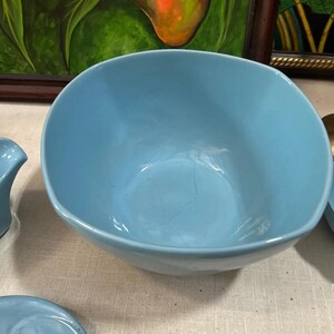 May include: A light blue ceramic bowl with a rounded square shape. The bowl has a smooth surface and visible cracks. The bowl is part of a set of light blue ceramic dishware.