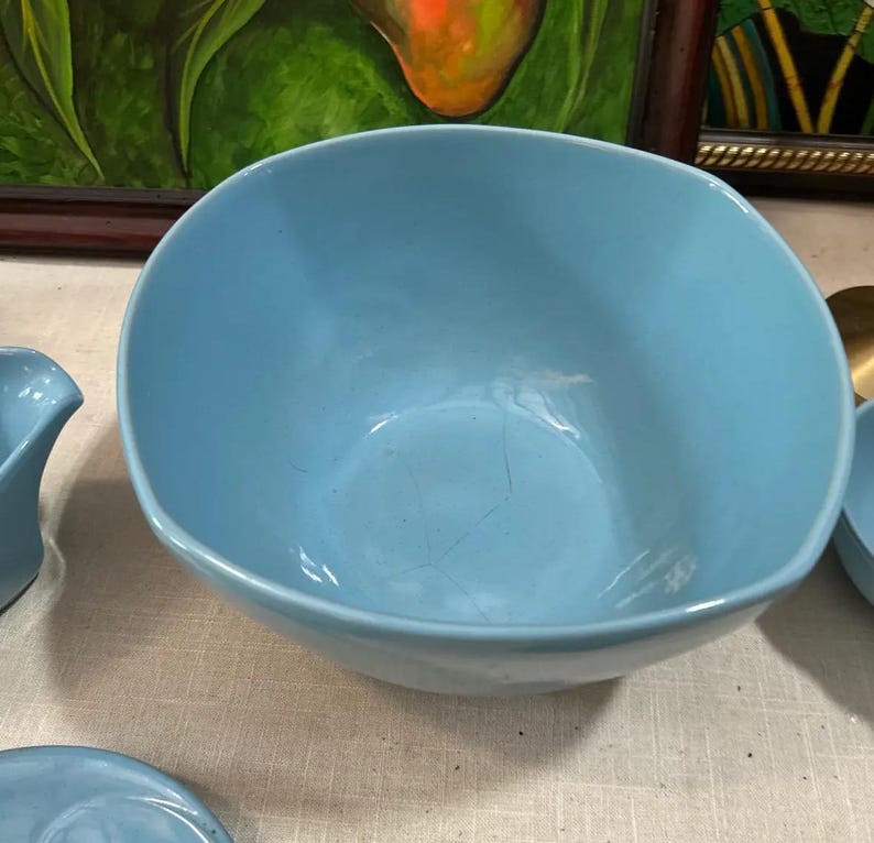 May include: A light blue ceramic bowl with a rounded square shape. The bowl has a smooth surface and visible cracks. The bowl is part of a set of light blue ceramic dishware.