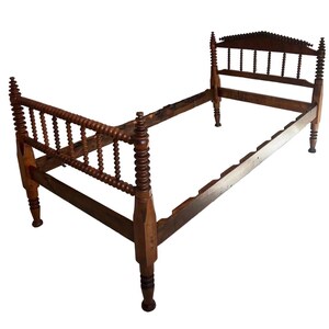May include: A dark brown wooden bed frame featuring a headboard and footboard with turned spindle detailing. The headboard has a decorative arched top rail with small carved points, and both ends are supported by four tall, cylindrical posts.