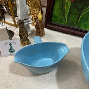 May include: A light blue ceramic gravy boat with a handle. The boat has a spout for easy pouring. A vintage Fig House sticker is visible in the background, along with other decorative items.