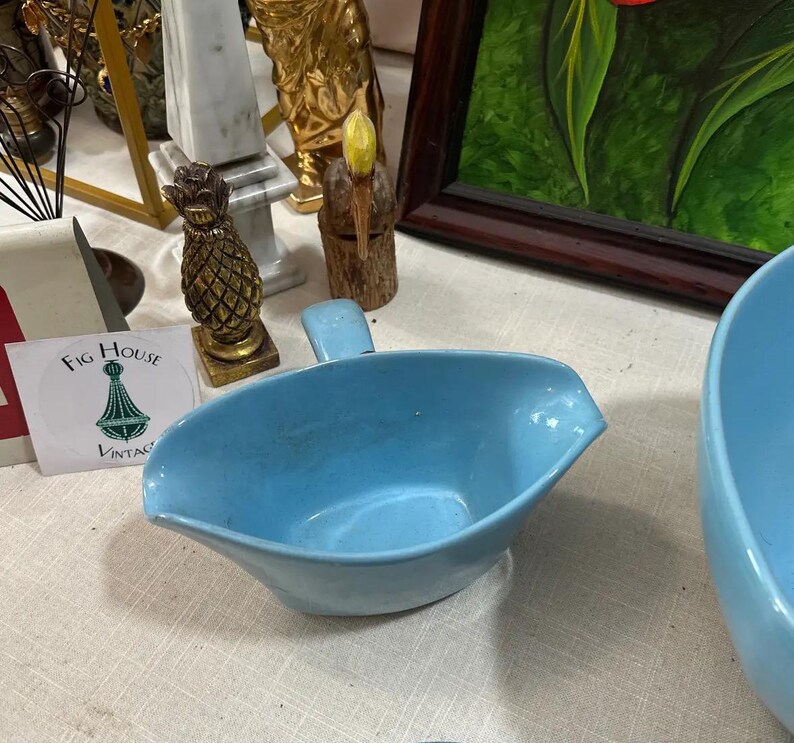 May include: A light blue ceramic gravy boat with a handle. The boat has a spout for easy pouring. A vintage Fig House sticker is visible in the background, along with other decorative items.