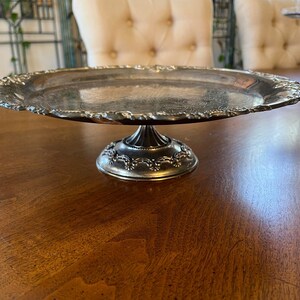 May include: A silver cake stand with a round, scalloped edge and a pedestal base. The stand is on a wooden table.