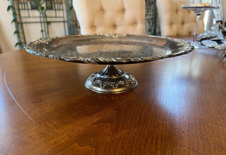 May include: A silver cake stand with a round, scalloped edge and a pedestal base. The stand is on a wooden table.