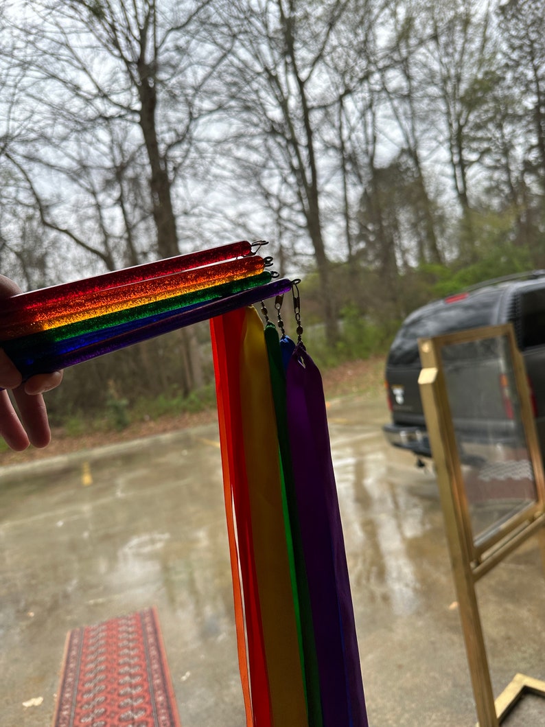 May include: A rainbow-colored streamer with multiple ribbons attached to a clear plastic wand. The ribbons are red, orange, yellow, green, blue, and purple.