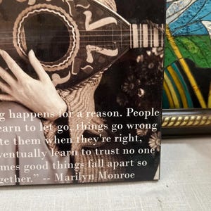 K&ouml;nnte beinhalten: Schwarz-Wei&szlig;-Bild mit einem Zitat von Marilyn Monroe: &bdquo;Alles geschieht aus einem Grund. Menschen lernen loszulassen, Dinge gehen schief...&ldquo; Das Zitat befindet sich auf einem Buchcover mit einer Gitarre und einer Hand.