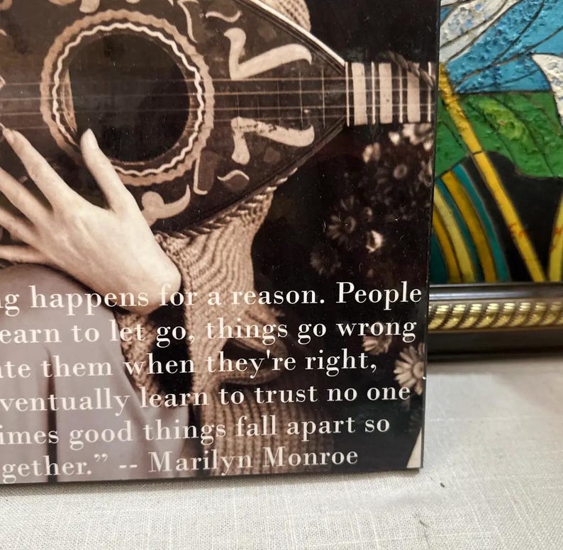 K&ouml;nnte beinhalten: Schwarz-Wei&szlig;-Bild mit einem Zitat von Marilyn Monroe: &bdquo;Alles geschieht aus einem Grund. Menschen lernen loszulassen, Dinge gehen schief...&ldquo; Das Zitat befindet sich auf einem Buchcover mit einer Gitarre und einer Hand.