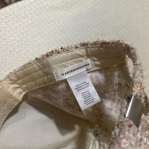 May include: A baseball cap with a textured, multi-colored design. The interior is a light cream color, with a tag that reads "by ANTHROPOLOGIE". The cap has a metal clasp.