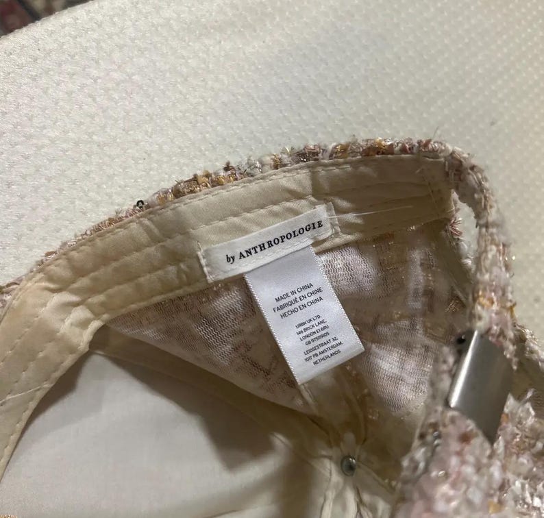 May include: A baseball cap with a textured, multi-colored design. The interior is a light cream color, with a tag that reads "by ANTHROPOLOGIE". The cap has a metal clasp.