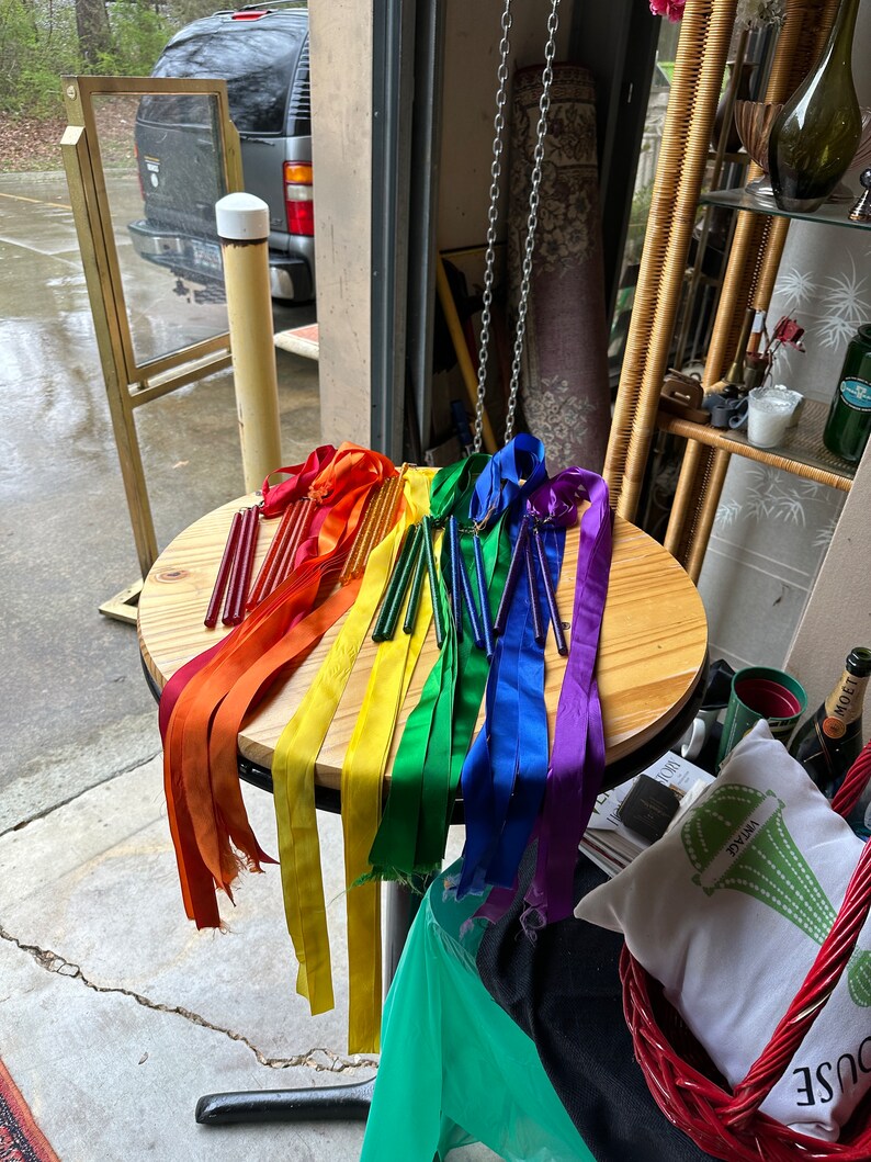 May include: A table with rainbow colored ribbons and wooden dowels. The ribbons are tied to the dowels and are hanging down. The table is made of wood and has a light brown finish.
