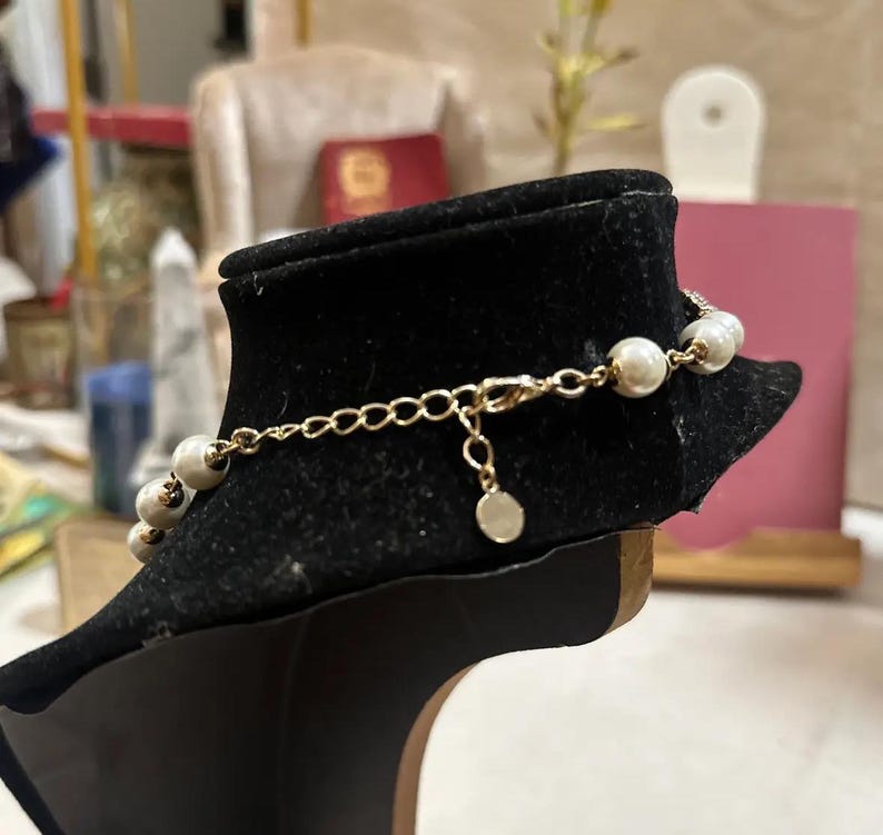Puede incluir: Una pulsera de cadena dorada con perlas de imitaci&oacute;n. La pulsera se exhibe en un expositor de joyer&iacute;a de terciopelo negro. Las perlas son blancas y est&aacute;n espaciadas uniformemente a lo largo de la cadena. El cierre y la cadena extensora son visibles.