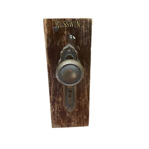 May include: A vintage brass door knob with a keyhole and a Russwin inscription on a wooden plaque.
