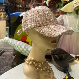 May include: A tweed baseball cap in shades of beige, tan, and white, displayed on a mannequin head. The cap has a curved brim and a textured appearance. A gold-toned pearl necklace adorns the mannequin's neck. Other hats and accessories are visible in the background.