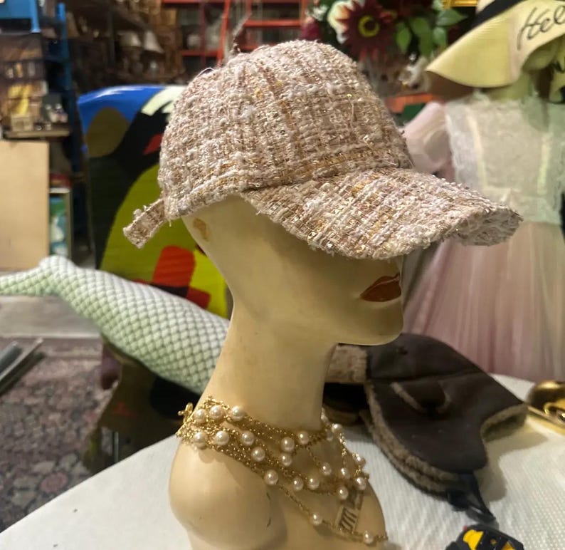May include: A tweed baseball cap in shades of beige, tan, and white, displayed on a mannequin head. The cap has a curved brim and a textured appearance. A gold-toned pearl necklace adorns the mannequin's neck. Other hats and accessories are visible in the background.