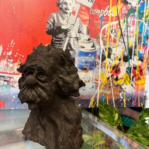 May include: A dark brown, textured, sculpted bust of a man with a long beard and a whimsical expression. The bust is made of a rough, earthy material and appears to be hand-crafted.