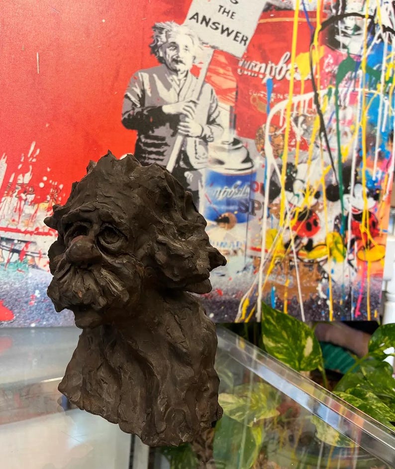 May include: A dark brown, textured, sculpted bust of a man with a long beard and a whimsical expression. The bust is made of a rough, earthy material and appears to be hand-crafted.