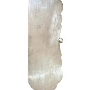 May include: A silver-colored, rectangular metal object with a scalloped edge on one side and a small, round knob. The metal has a brushed texture, and the object appears to be a decorative or functional item.