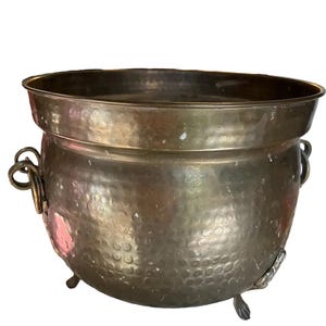 May include: A vintage bronze cauldron with a hammered texture. The cauldron has a wide rim, two handles, and three decorative feet. The metal has a tarnished appearance, suggesting age and use. This decorative piece could be used for display or storage.