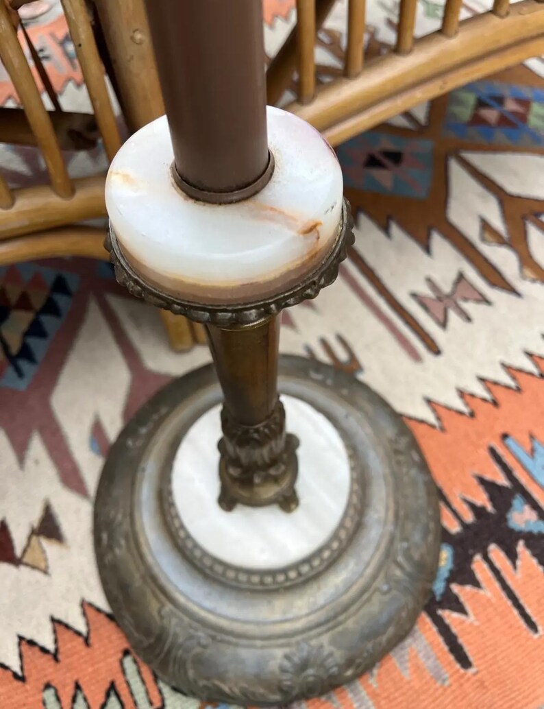 Puede incluir: Una l&aacute;mpara de pie antigua con un poste marr&oacute;n y una base de m&aacute;rmol blanco. La l&aacute;mpara presenta una base de metal decorativa con un dise&ntilde;o ornamentado. El dise&ntilde;o de la l&aacute;mpara es vintage y elegante.