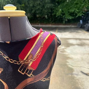 May include: A black sleeveless dress with a colorful chain and buckle print. The dress has a scoop neck and a button detail on the shoulder.