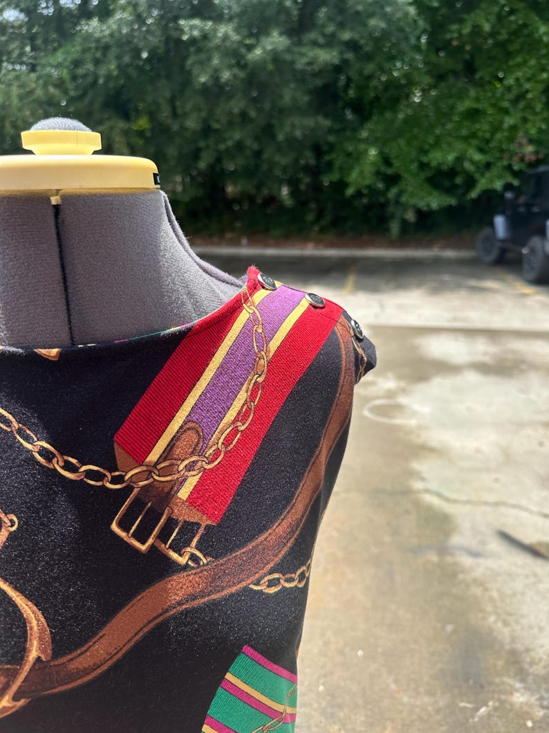 May include: A black sleeveless dress with a colorful chain and buckle print. The dress has a scoop neck and a button detail on the shoulder.