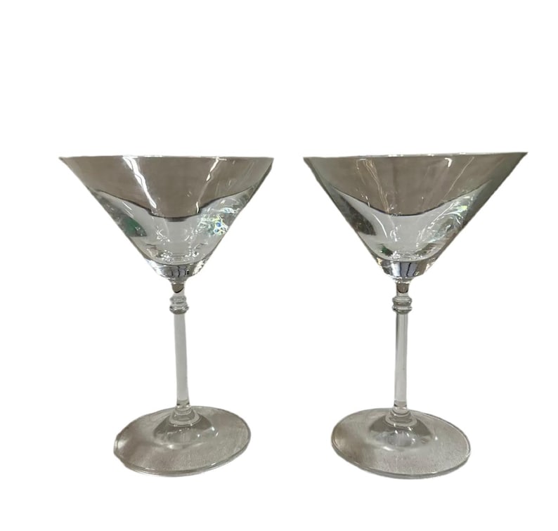 May include: A pair of clear martini glasses with silver rims. Each glass features a triangular bowl, a long stem, and a round base. The glasses are empty and presented against a white backdrop. The design is classic and elegant.