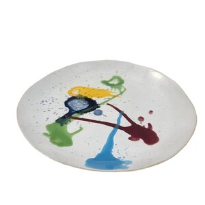 Op de afbeelding: Witte keramische plaat met kleurrijke abstracte verfspatten in geel, blauw, groen en bordeaux. De plaat heeft een gespikkelde oppervlakte en een enigszins onregelmatige vorm.