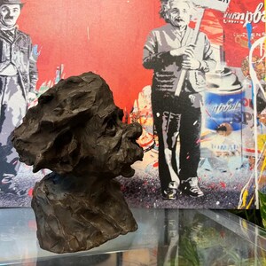 May include: A bronze sculpture of a person's head and shoulders. The sculpture is in a realistic style and has a textured surface. The sculpture is on a glass surface in front of a colourful painting.