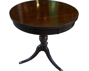 May include: A round, dark brown wooden pedestal table with a smooth, polished surface. The table features a central column support that branches into three curved legs, each ending in a small, metallic, claw-shaped foot detail.